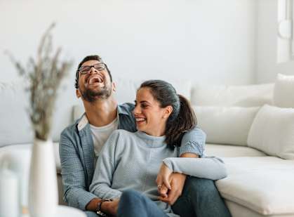 A man and a woman are sitting on a white couch, laughing together.