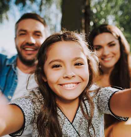 A young girl is taking a selfie with two adults in a park.