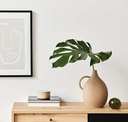 A framed artwork hangs on a wall above a desk with a chair, a plant, and some books.