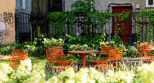 A patio with orange chairs and tables is surrounded by green plants.
