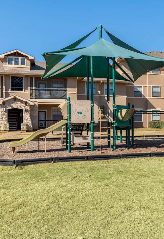 A playground with a green canopy and a yellow slide is in front of apartment buildings.