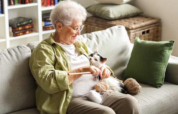 An elderly woman is knitting while sitting on a couch with a cat nearby.