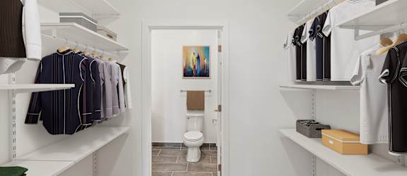 A white walk in closet with clothes hanging and a toilet in the background.