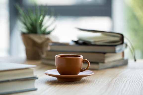 A cup of coffee sits on a saucer in front of a stack of books and a pair of glasses on a wooden table.