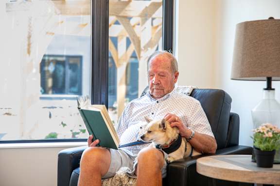 an older man sits in a chair with his dog and reads a book