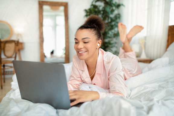 A woman in a pink robe is lying on a bed using a laptop.