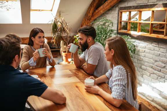 A group of people are sitting around a wooden table, each holding a cup.