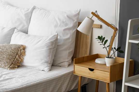 A bed with white pillows and a wooden nightstand with a lamp and plant.
