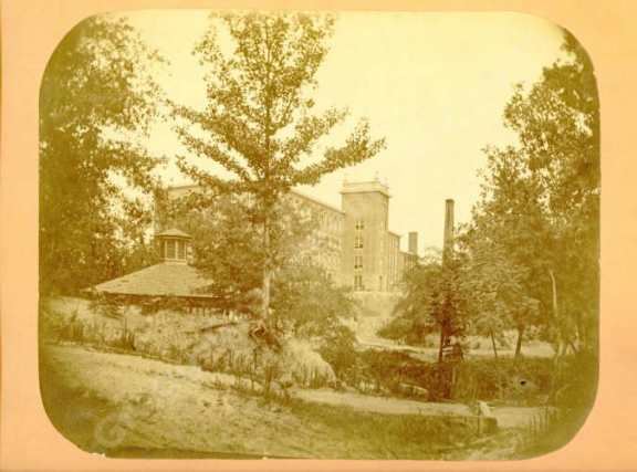 A sepia-toned photograph of a house with a large tree in front and a building in the background.
