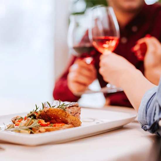 A person is holding a glass of red wine while sitting at a table with a plate of food in front of them.