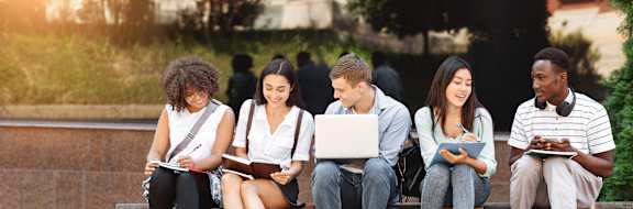 A group of five students are sitting on a bench outside, each using a different device to study or work.