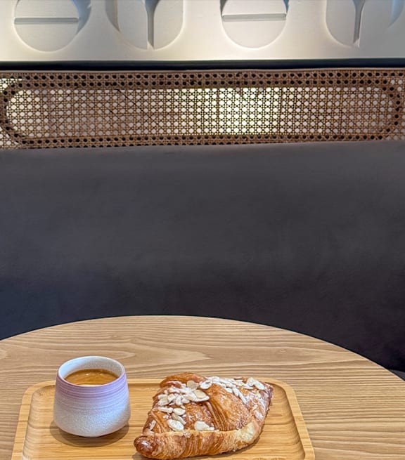 A pastry with almonds on top is on a wooden board with a cup of coffee.