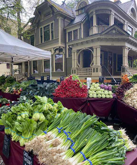 A market with vegetables on display in front of a Victorian style house.