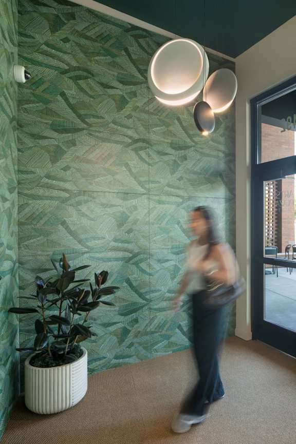 A woman walks past a green wall and a plant in a white pot.