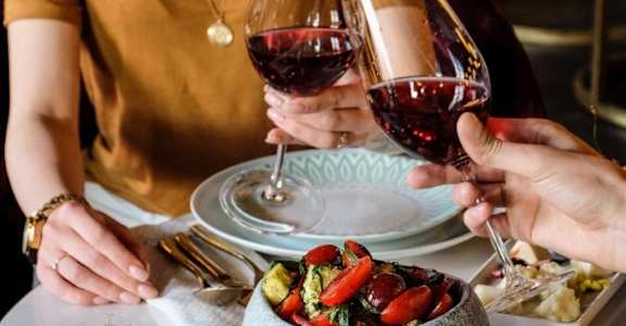 A woman is toasting with a glass of red wine at a table with a variety of foods.