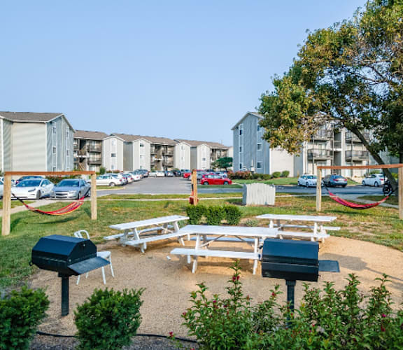 A park area with picnic tables and BBQ grills in front of apartment buildings.