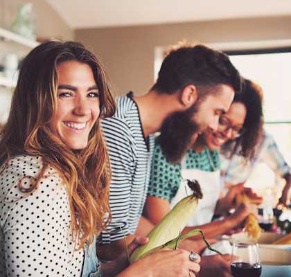 A woman with a polka dot shirt is holding a corn cob while two people prepare food in the background.
