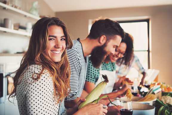 A woman with a polka dot shirt is holding a corn cob while two people prepare food in the background.