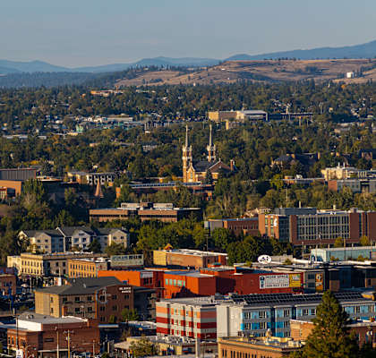 A cityscape with buildings of various sizes and colors, surrounded by trees and hills in the distance.