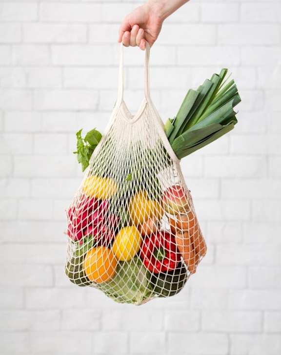 A hand holding a mesh bag filled with various fruits and vegetables.
