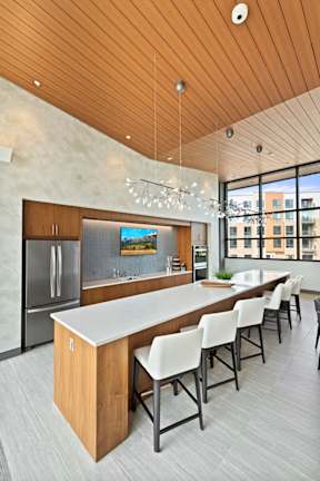 A community kitchen with a long quartz island, bar stool seating, a sink, refrigerator, a large window, and modern pendant chandeliers. at Keva Flats, Exton, PA