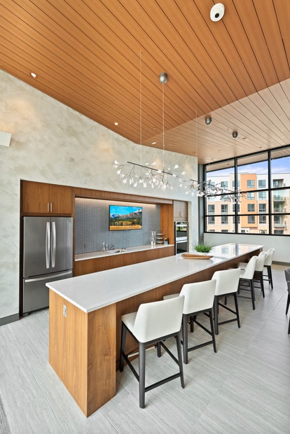 A community kitchen with a long quartz island, bar stool seating, a sink, refrigerator, a large window, and modern pendant chandeliers. at Keva Flats, Exton, PA