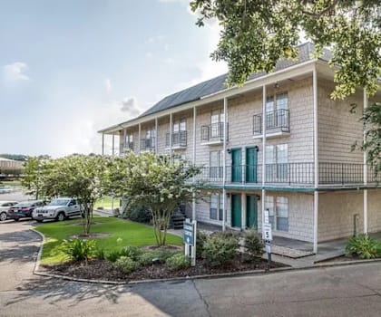 A building with a balcony and a green door is surrounded by trees and has a parking lot in front.