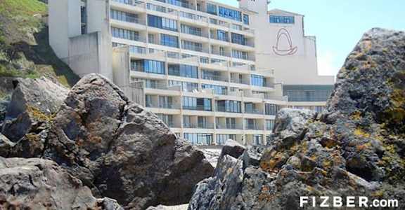 A large white building with a red logo on the side is situated on a rocky hillside.