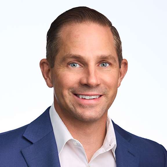 A man in a dark suit and white shirt smiles at the camera.