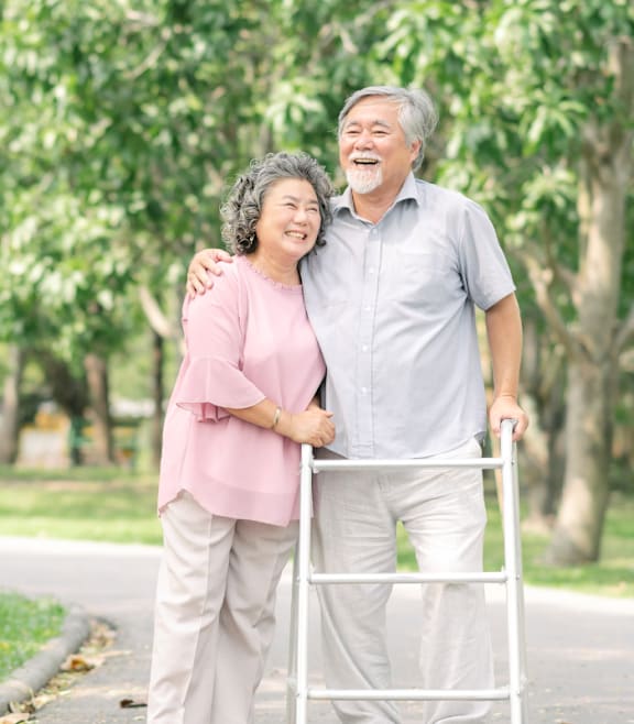 An elderly couple is standing in a park with the woman holding a walker.