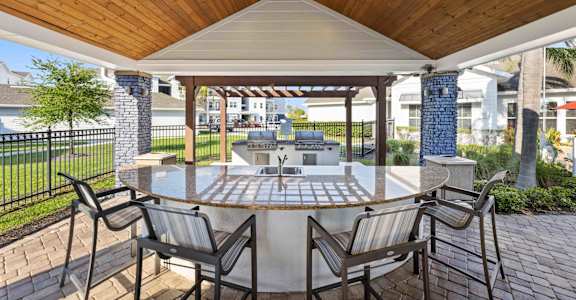 a patio with a large circular table and chairs with a grill in the background