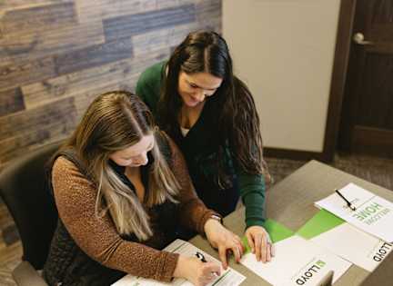 Two women are sitting at a table, looking at papers and writing on them.
