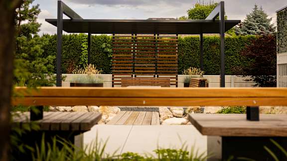A wooden bench is in the foreground of a park with a wooden pergola above it.