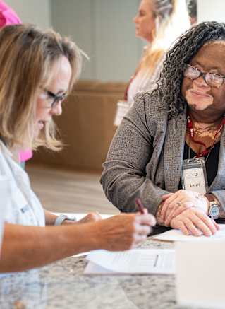 Two women are sitting at a table, one is writing on a piece of paper while the other looks on.