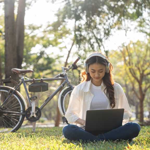 A person is sitting on the grass with a laptop and a bicycle is parked behind them.