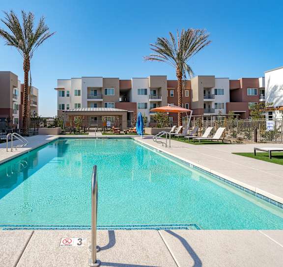A swimming pool in front of apartment buildings with sun loungers on the side.