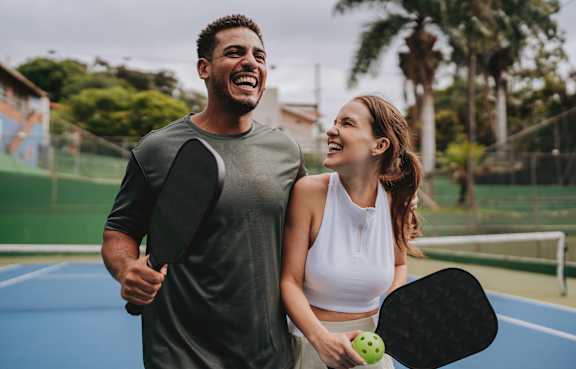 A man and a woman are standing on a tennis court, both holding tennis rackets and smiling.