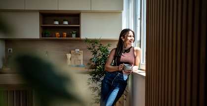 A woman in a brown top and blue jeans stands by a window in a modern kitchen.