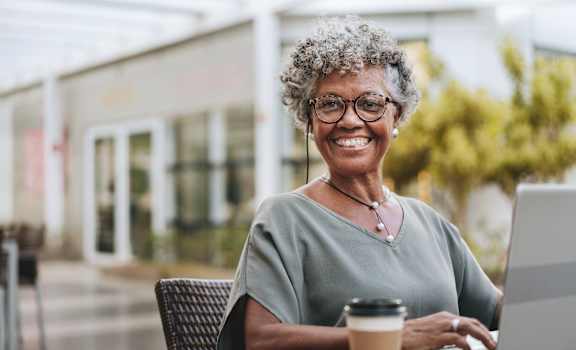 A woman with glasses and a grey top is smiling while using a laptop and holding a cup of coffee.