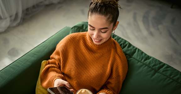 A woman in an orange sweater is sitting on a green couch, holding a cup of coffee and looking at her phone.