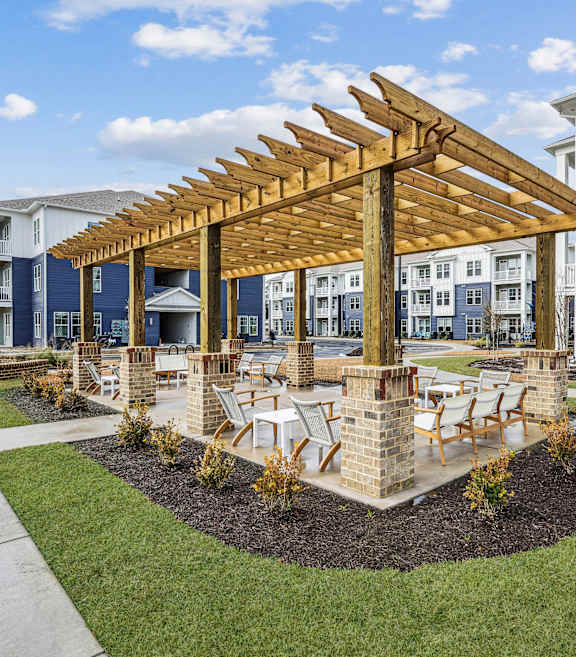 A wooden pavilion is in the middle of a grassy area with a building in the background.