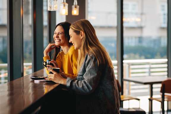 Two women are sitting at a table, one is holding a phone and the other is holding a cup.