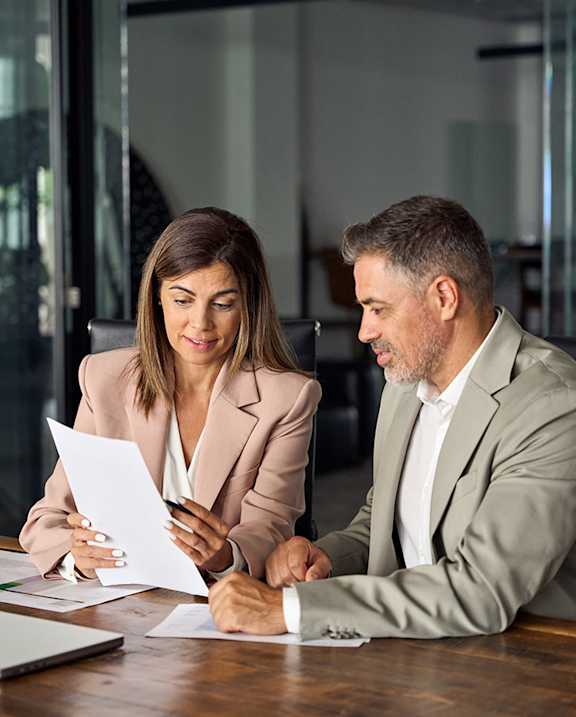Two people in suits are sitting at a table with papers, appearing to have a serious discussion.