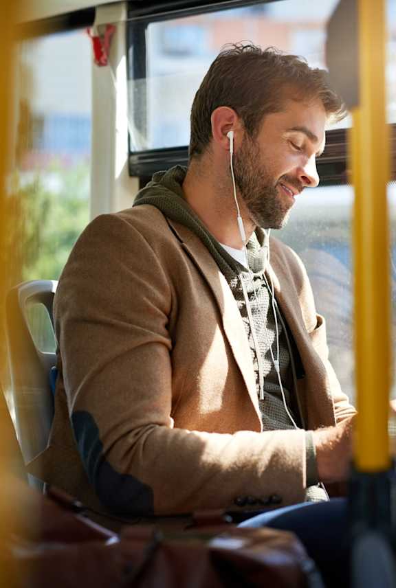 A man is sitting on a bus, wearing a brown jacket and holding a black tablet.