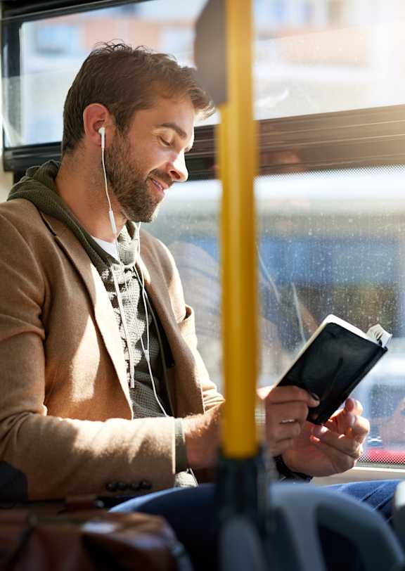 A man is sitting on a bus, wearing a brown jacket and holding a black tablet.
