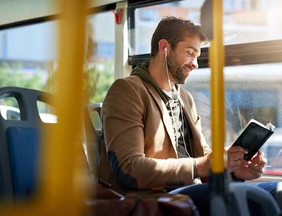 A man is sitting on a bus, wearing a brown jacket and holding a black tablet.
