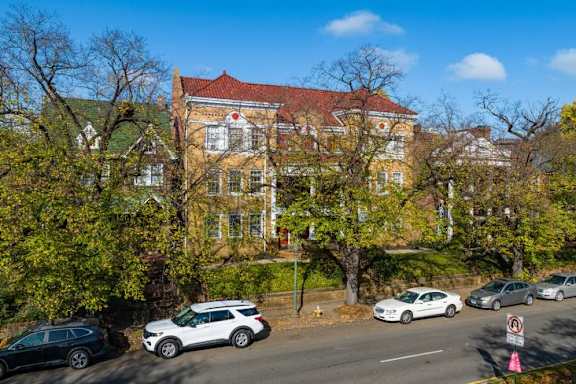 A red-roofed building is surrounded by trees and cars are parked on the street.