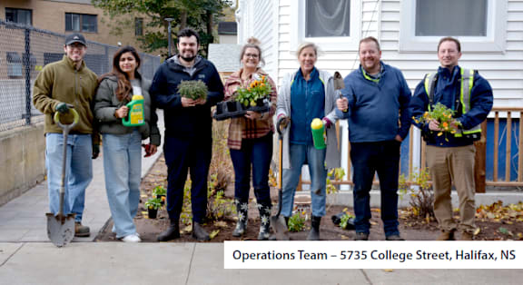 A group of people standing on a sidewalk holding plants and gardening tools.