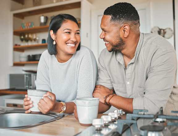A man and a woman are sitting at a kitchen counter, both holding mugs and smiling at each other.
