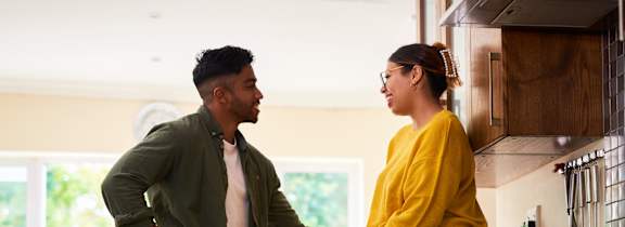 A man and woman are standing in a kitchen, with the woman sitting on the counter and the man standing next to her.